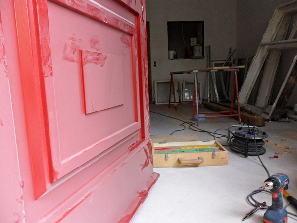 Ein Raum in Renovierung mit einer rot grundierten Tür im Vordergrund. Im Hintergrund sind ein Tisch, verschiedene Werkzeuge, eine Kabeltrommel und Fensterrahmen zu sehen. Ein blauer Bohrer liegt auf dem Boden.