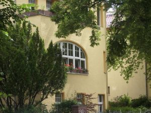 Gelbes Gebäude mit einem abgerundeten Fenster, umgeben von grünen Bäumen und Büschen. Auf der Fensterbank stehen blühende Blumenkästen. Der Großteil des Gebäudes wird von sattem Laub und einem kleinen Garten eingeschlossen.