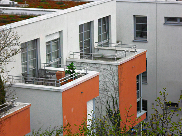Mehrfamilienhaus mit hellgrauer und orangefarbener Fassade. Zwei Balkone mit weißen Geländern sind zu sehen, einer hat eine Topfpflanze und Gartenmöbel. Im Vordergrund sind Äste von Bäumen.