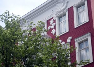 Fassadenansicht eines Gebäudes mit roten Backsteinen und weißen Stuckverzierungen. Der obere Teil zeigt florale Reliefs. Vor dem Gebäude steht ein Baum mit grünen Blättern, teilweise die Sicht auf die Fenster verdeckend.