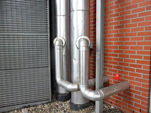 Silberne Rohre und Lüftungsschächte an einer Ziegelmauer. Die Rohre erstrecken sich vertikal und sind miteinander verbunden. Rechts daneben befindet sich ein Metallzaun. Der Boden ist mit Kies bedeckt.