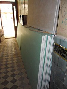 Stapel von Gipskartonplatten lehnen an einer gefliesten Wand in einem schmalen Flur mit kariertem Boden. Im Hintergrund ist eine Tür mit Lichtdurchschein zu sehen. Links der Platten befinden sich einige kleinere, aufgestapelte Gegenstände.