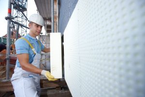 Ein Bauarbeiter mit Schutzhelm und Handschuhen befestigt eine Dämmplatte an einer Außenwand eines Gebäudes. Im Hintergrund ist ein Baugerüst zu sehen. Der Arbeiter trägt blaue Kleidung und weiße Latzhose.