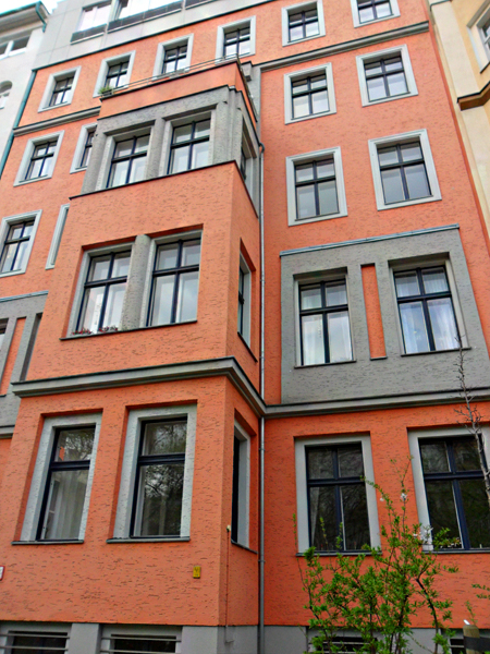 Alt-Text: Ein mehrstöckiges Gebäude mit orangefarbener Fassade, grauen Fensterumrandungen und mehreren weißen Fenstern. Einige Fensterbänke haben kleine Vorsprünge. Vor dem Gebäude sind Pflanzen und ein Baum zu sehen.