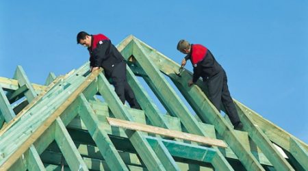Zwei Dachdecker in Arbeitskleidung montieren bei blauem Himmel Holzbalken auf einem neu errichteten Dachstuhl aus grün imprägniertem Holz.