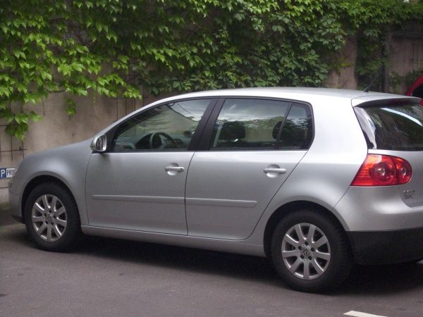 Silberner Kompaktwagen, geparkt auf einem Parkplatz vor einer mit Efeu bewachsenen Wand. Das Auto hat fünf Türen und die Rückleuchten sind rot. Der Asphaltboden ist sichtbar, und das Fahrzeug steht in einem schattigen Bereich.