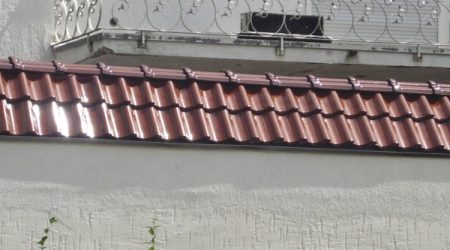 Hohlziegel aus glänzendem Material auf einem kleinen geneigten Dachabschnitt vor einer weißen Fassade mit dekorativem Balkon aus Schmiedeeisen.