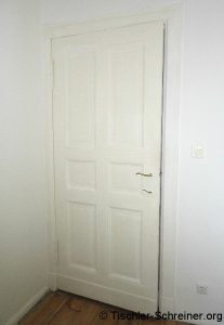Weiße Holztür mit klassischem Kassettenprofil in einem Innenraum, inklusive Messinggriff und umlaufendem Türrahmen; daneben ein Lichtschalter und eine Steckdose.