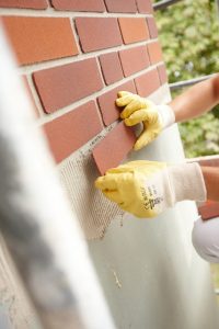 Eine Person setzt mit gelben Schutzhandschuhen eine Ziegelplatte an eine Wand. Die Wand ist teils mit parallelen roten Ziegelplatten bedeckt, und es sind Baustellengerüste im Hintergrund zu sehen.