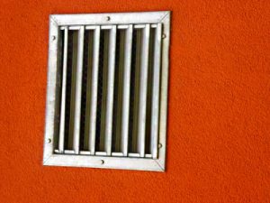 Ein rechteckiges Metallgitter mit sieben vertikalen Stäben ist in eine leuchtend orangefarbene Wand eingelassen.