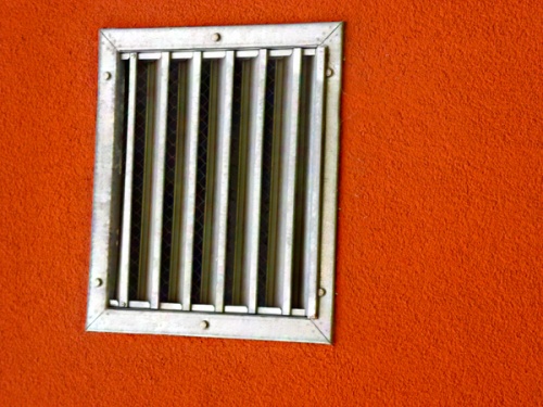 Ein rechteckiges Metallgitter mit sieben vertikalen Stäben ist in eine leuchtend orangefarbene Wand eingelassen.