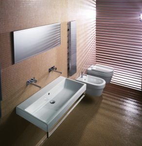Modernes Badezimmer mit rechteckigem Waschbecken, an der Wand montierten Armaturen und großem Spiegel. Auf der rechten Seite befinden sich eine Toilette und ein Bidet. Die Wände sind mit Mosaikfliesen verkleidet, und eine Lamellenstruktur sorgt für Lichteffekte.