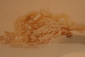Wasserfleck an der Zimmerdecke mit unregelmäßigen braunen Verfärbungen, die auf einen möglichen Wasserschaden hinweisen. Der Fleck erstreckt sich über einen großen Bereich und hat eine amorphe Form, die die helle Wandfarbe überlagert.