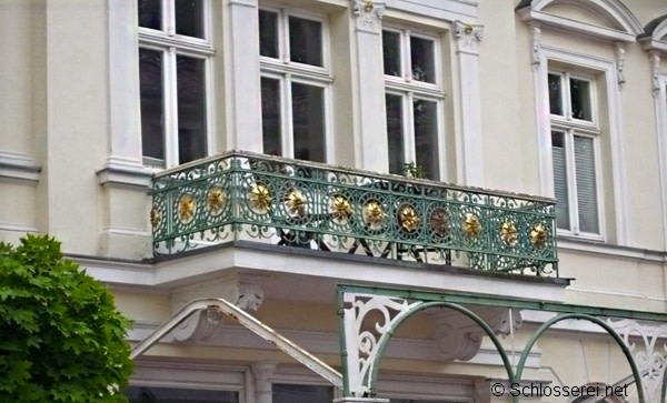 Ein eleganter Balkon mit kunstvollen, grün-goldenen Eisenornamenten an einem historischen Gebäude. Das Geländer zeigt detaillierte Verzierungen mit floralen Mustern. Im Hintergrund sind weiße Fenster und ein Teil der Fassade zu sehen.