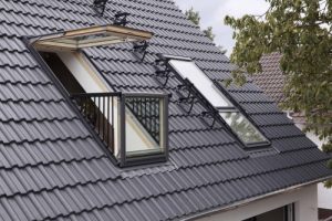 Ein geöffneter Dachbalkon mit zwei großen Dachfenstern in einem dunklen Ziegeldach. Das untere Fenster ist senkrecht aufgestellt und mit einem schmalen Geländer versehen, das obere Fenster ist nach außen oben geöffnet.