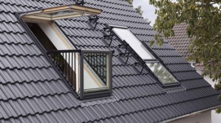Ein geöffneter Dachbalkon mit zwei großen Dachfenstern in einem dunklen Ziegeldach. Das untere Fenster ist senkrecht aufgestellt und mit einem schmalen Geländer versehen, das obere Fenster ist nach außen oben geöffnet.