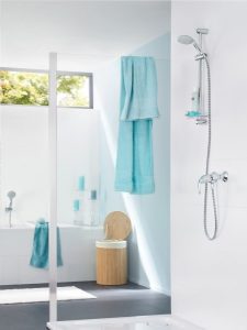 Moderne, helle Badezimmer mit Dusche rechts und Badewanne links. Türkisfarbene Handtücher hängen an der Dusche und nahe der Badewanne. Ein Holzhocker steht zwischen Dusche und Badewanne. Tageslicht strömt durch ein großes Fenster.