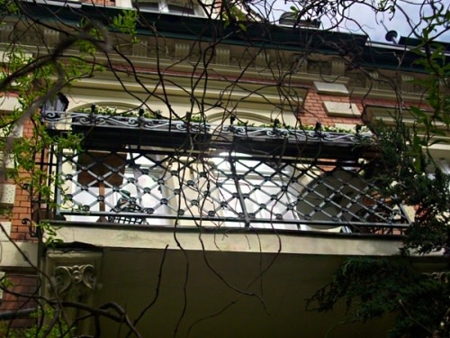 Ein Balkon eines Backsteingebäudes mit kunstvollem Metallgeländer. Kletterpflanzen ranken um das Geländer. Durch die Äste schimmert Sonnenlicht. Die Fassade zeigt architektonische Verzierungen.