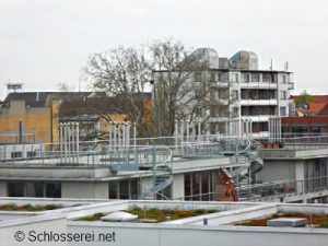 Blick auf ein modernes Gebäude mit einer Wendeltreppe auf dem Dach, umgeben von weiteren Gebäuden. Im Hintergrund sind kahle Bäume sichtbar. Der Himmel ist bewölkt. Im Vordergrund sieht man ein begrüntes Flachdach.