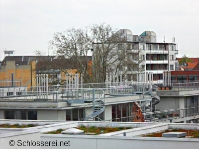 Blick auf ein modernes Gebäude mit einer Wendeltreppe auf dem Dach, umgeben von weiteren Gebäuden. Im Hintergrund sind kahle Bäume sichtbar. Der Himmel ist bewölkt. Im Vordergrund sieht man ein begrüntes Flachdach.