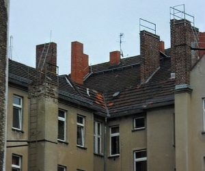 Mehrstöckige Altbauten mit teils sanierungsbedürftigen Fassaden und zahlreichen Schornsteinen auf den Ziegeldächern. Einige Kamine haben Metalltritte und Steigleitern für den Schornsteinfeger. Das Wetter ist bewölkt, die Szene wirkt urban und dicht bebaut.