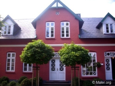 Rotes Backsteinhaus mit dunklem Schieferdach. Drei weiße Gaubenfenster im Dach. Zwei Bäume mit kugelförmigen Kronen vor dem Eingang. Weiße Türen und Fenster mit Sprossendekor und Blumenschmuck. Klarer Himmel im Hintergrund.