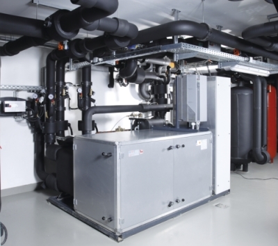 Industriekühlsystem in einem technischen Raum. Zu sehen sind zahlreiche Rohre, ein großer grauer Kasten und verschiedene Anschlüsse. Die Umgebung wirkt sauber und ordentlich installiert, mit schwarzer Isolierung auf den Rohren.