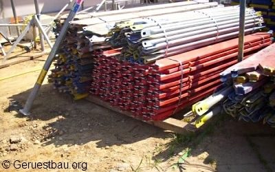 Gestapelte Gerüstbohlen und Vertikalrahmen aus Metall auf einer Baustelle, bereit zur Montage eines Fassadengerüsts für Bau- oder Sanierungsarbeiten.