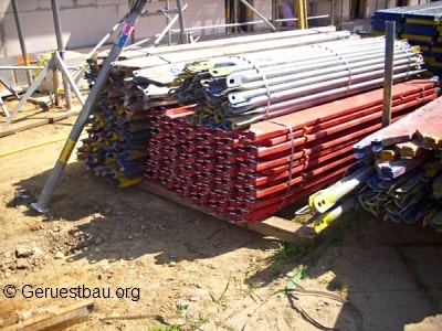 Gestapelte Gerüstbohlen und Vertikalrahmen aus Metall auf einer Baustelle, bereit zur Montage eines Fassadengerüsts für Bau- oder Sanierungsarbeiten.
