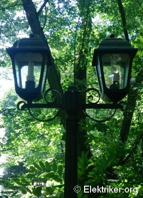 Zwei klassische Laternen an einem dunklen Metallmast stehen vor einem Hintergrund aus dichtem, grünem Laub und Ästen. Die Umgebung wirkt ruhig und naturverbunden. Rechts unten im Bild ist der Text "© Elektriker.org" zu sehen.