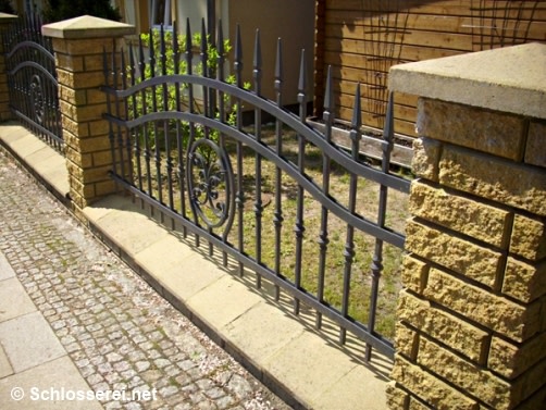 Ein elegant geschwungenes Metallzaun mit dekorativen Spitzen und einem runden Ornament in der Mitte. Der Zaun ist auf einem niedrigen gelben Steinfundament montiert, das entlang eines Gehwegs verläuft. Im Hintergrund sind Büsche und ein Holzzaun zu sehen.