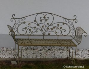 Eine elegante schmiedeeiserne Gartenbank mit geschwungenen Verzierungen steht vor einer weißen Hauswand. Der Boden ist mit weißen Steinen bedeckt. Im Vordergrund ist ein schmaler Rasenstreifen zu sehen.