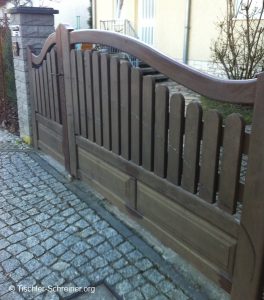 Großes zweiflügeliges Holztor mit geschwungener Oberkante und senkrechten Latten, eingebettet in eine gepflasterte Einfahrt.