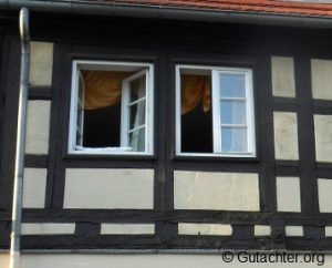 Fachwerkhaus mit zwei geöffneten Fenstern im Obergeschoss. Die Fensterscheiben sind klar und ein gelber Vorhang ist zu sehen. Die Hausfassade zeigt das typische Muster aus dunklen und hellen Holzbalken.