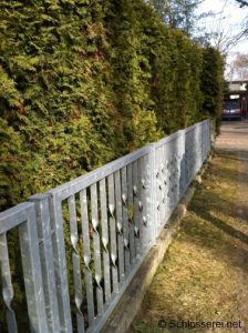 Metallzaun mit Rautenmuster entlang einer hohen, dichten Hecke aus Koniferen. Die Sonneneinstrahlung erzeugt Schatten auf dem Rasen und dem Gehweg. Im Hintergrund sind Bäume und ein geparktes Auto sichtbar.