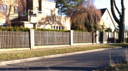 Langer, klassischer Holzzaun mit senkrechten Latten und stabilen Betonpfosten, der mehrere Grundstücke an einer Wohnstraße abgrenzt.