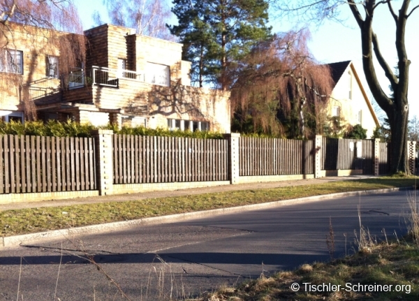 Langer, klassischer Holzzaun mit senkrechten Latten und stabilen Betonpfosten, der mehrere Grundstücke an einer Wohnstraße abgrenzt.