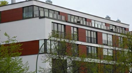Modernes Mehrfamilienhaus mit Flachdach, großzügigen Fensterfronten und einer Fassade aus weißen und rotbraunen Paneelen, teilweise von Bäumen verdeckt.