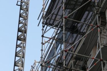 Stahlrahmengerüst mit Auslegern und Fangschutznetzen an einer mehrstöckigen Fassade, neben einem Baukran bei Arbeiten in großer Höhe.
