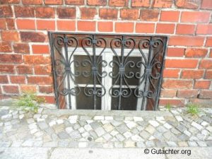 Ein kleines Fenster eines Backsteingebäudes ist mit einem kunstvollen schwarzen Metallgitter gesichert. Davor liegen unregelmäßig gepflasterte Steine mit vereinzeltem Grasbewuchs.