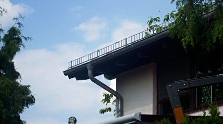 Hausdach mit Schneefanggitter: Ein modernes Steildach mit Dachrinne, Fallrohr und aufgesetztem Schneefanggitter entlang der Dachkante. Im Vordergrund befinden sich Bäume und ein Sonnenschutzgestänge.