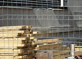 Gestapeltes Bauholz auf einer Baustelle hinter einem Drahtzaun. Im Hintergrund ist die Fassade eines Gebäudes mit Lüftungsschlitzen zu sehen. Das Holz ist ordentlich aufgeschichtet und bereit zur weiteren Verwendung im Bauprozess.