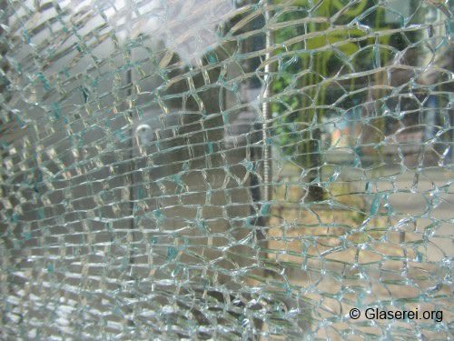 Zersprungenes Sicherheitsglas mit feinem Netz aus Rissen. Im Hintergrund verschwommenes Bild einer grünen Umgebung. Das Glas ist in Gitterformen gesprungen und zeigt die Struktur eines möglichen Bruchmusters.
