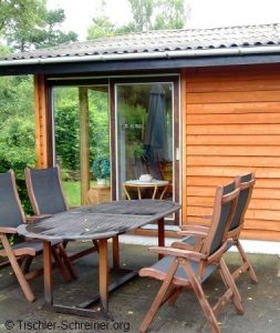 Gartenmöbel-Set mit Holztisch und sechs Klappstühlen auf einer Terrasse vor einem Holzhaus mit Schiebetür.