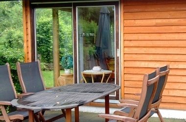 Gartenmöbel-Set mit Holztisch und sechs Klappstühlen auf einer Terrasse vor einem Holzhaus mit Schiebetür.