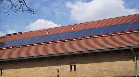 Ziegeldach eines historischen Gebäudes mit einer langen Reihe flacher Solarthermie-Kollektoren. Die Kollektoren sind parallel zur Dachfläche montiert. Das Gebäude ist aus gelbem Klinker mit roten Zierbögen über den Fenstern gebaut.