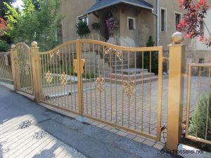 Ein elegantes, goldenes schmiedeeisernes Tor mit dekorativen Ornamenten steht vor einem gepflegten Haus mit Garten. Das Tor ist geschlossen und umrahmt einen gepflasterten Weg, der zu einer Treppe und einer Eingangstür führt.