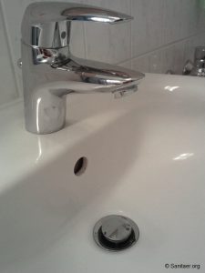 Ein Waschbecken mit einem glänzenden, verchromten Wasserhahn. Der Abfluss hat einen Stopfen und es gibt eine kleine Öffnung für Überlauf. Die Wand im Hintergrund ist mit weißen Fliesen verkleidet.