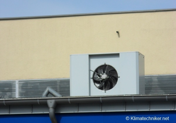Ein großer Außenventilator ist auf dem Dach eines Gebäudes angebracht. Der Ventilator ist in einem rechteckigen Gehäuse an der Wand befestigt. Im Hintergrund sieht man die beige Fassade des Gebäudes und den blauen Himmel.