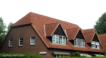 Mehrfamilienhaus mit rotem Ziegeldach in Deutschland. Das Dach weist mehrere gleichmäßig angeordnete Gauben mit dunkler Holzverkleidung und weißen Fenstern auf. Die Fassade besteht aus rotem Klinkermauerwerk, umgeben von dichter Begrünung.